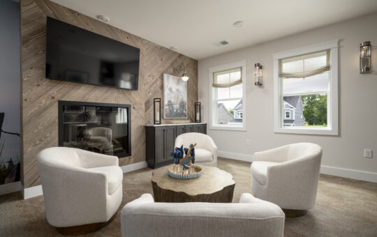 A modern living room with four white armchairs around a wooden coffee table, a wall-mounted TV above a fireplace, large windows with roman shades, and neutral decor.