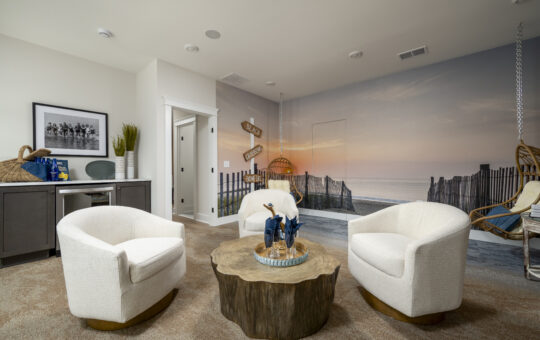 A cozy sitting area with two white swivel chairs and a wooden coffee table. The wall features a beach mural with a sunset, sand dunes, and a fence. To the left is a built-in desk with a chair and beach-themed decor.