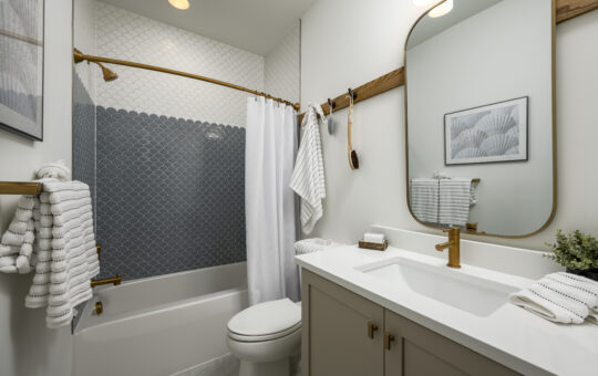 Modern bathroom with white walls, grey vanity, gold fixtures, and a large mirror. The bathtub has a patterned dark tile accent and a white shower curtain. Towels and decor complete the clean, stylish look.