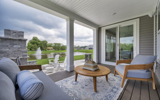 A cozy covered patio with cushioned chairs, a round wooden table, and a blue-patterned rug overlooks a pond with a fountain, green lawn, and white Adirondack chairs under a cloudy sky.