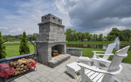 A stone outdoor fireplace on a patio with white Adirondack chairs nearby, firewood stacked to the left, and a pond with a fountain and trees in the background under a partly cloudy sky.