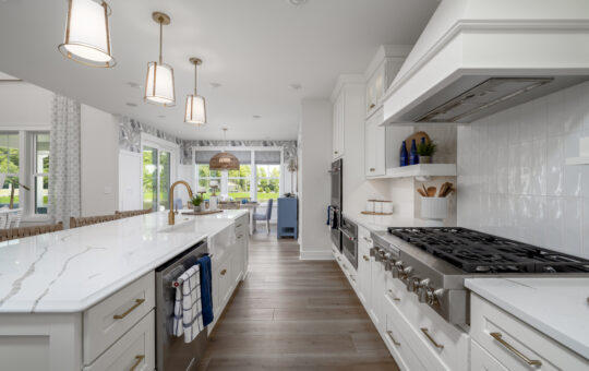 Modern kitchen with white cabinets, marble countertops, stainless steel appliances, a gas stove, pendant lights, and wood flooring. Large windows and a dining area with blue chairs are visible in the background.