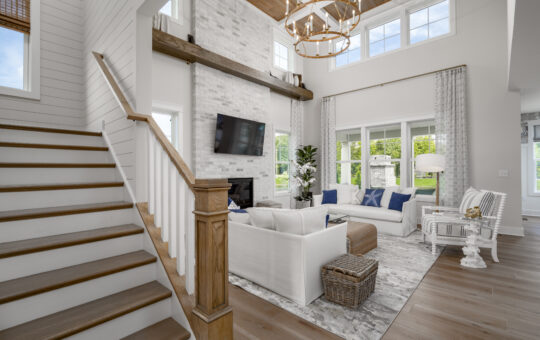 A bright, airy living room with high ceilings, large windows, and neutral décor. There’s a staircase with wooden railings, white sofas, a rug, TV above a fireplace, and modern chandeliers. Greenery is visible outside.