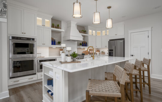 Modern kitchen with white cabinets, stainless steel appliances, a large marble island with a gold faucet, and four woven bar stools; pendant lights hang above the island, and decorative items are on the counters and shelves.