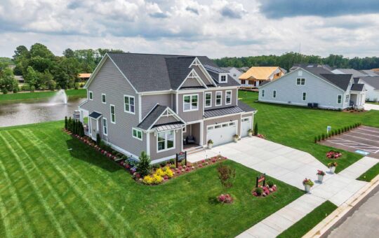 A two-story suburban house with gray siding, white trim, and a two-car garage sits on a manicured lawn with colorful flower beds. Theres a pond with a fountain behind the house and other similar homes nearby.