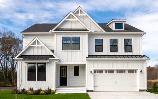 A modern two-story white house with black trim, large windows, a covered front porch, and a double garage, sits on a green lawn with a clear blue sky and trees in the background.