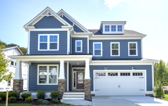 A modern two-story house with blue siding, white trim, a covered front porch, stone pillars, and a double garage door. Well-kept lawn and shrubs border the walkway leading to the entrance.