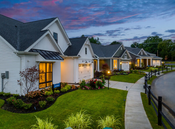 A row of modern, single-story suburban homes by Luxury Living by Foxlane Homes features well-manicured lawns and gardens, illuminated at dusk under a colorful sky. A curved sidewalk and black fence elegantly line the street.