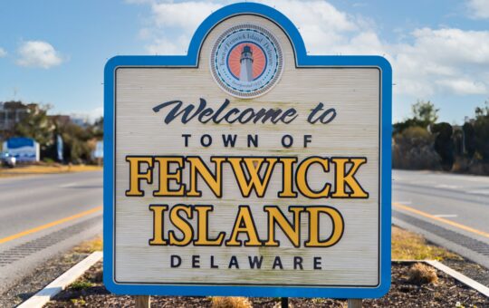 A wooden sign by the roadside reads Welcome to Town of Fenwick Island, Delaware with a lighthouse logo at the top, under a blue sky with scattered clouds.