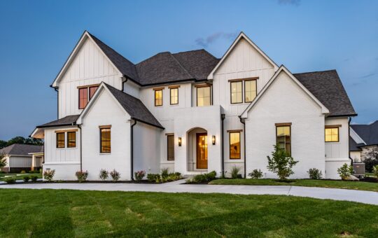 A large, modern two-story house with white brick and siding exterior, multiple peaked roofs, and warmly lit windows, surrounded by a manicured lawn and small shrubs at dusk.