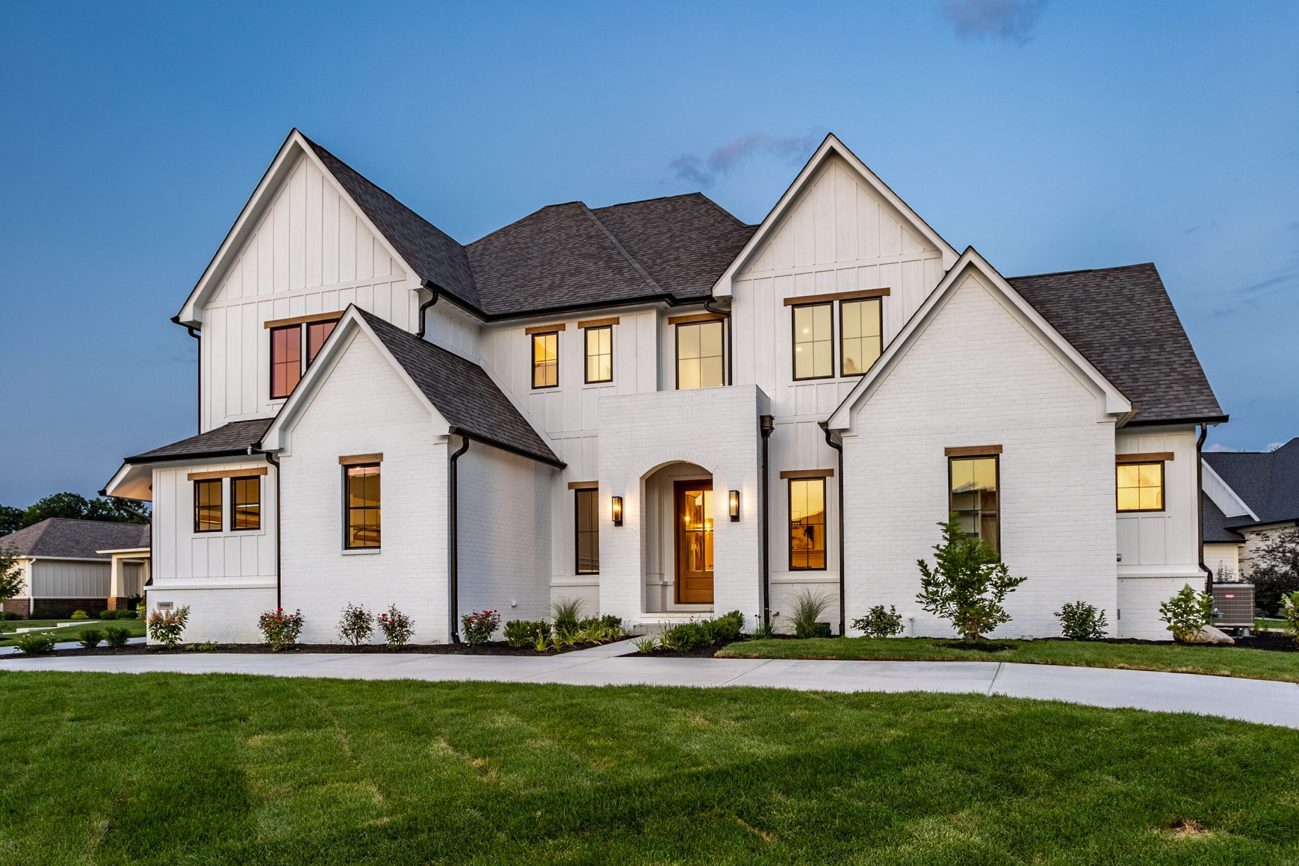 A large, modern two-story house with white brick and siding exterior, multiple peaked roofs, and warmly lit windows, surrounded by a manicured lawn and small shrubs at dusk.