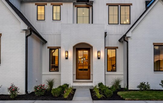 Modern white brick house with wood trim, large windows, a central wooden front door under an arched entryway, black sconces, and landscaped garden beds with shrubs and flowers along a concrete path.