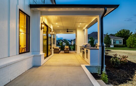 A modern covered patio at dusk, featuring outdoor seating and a built-in grill, with warm lighting and a view of a green lawn and neighboring houses.