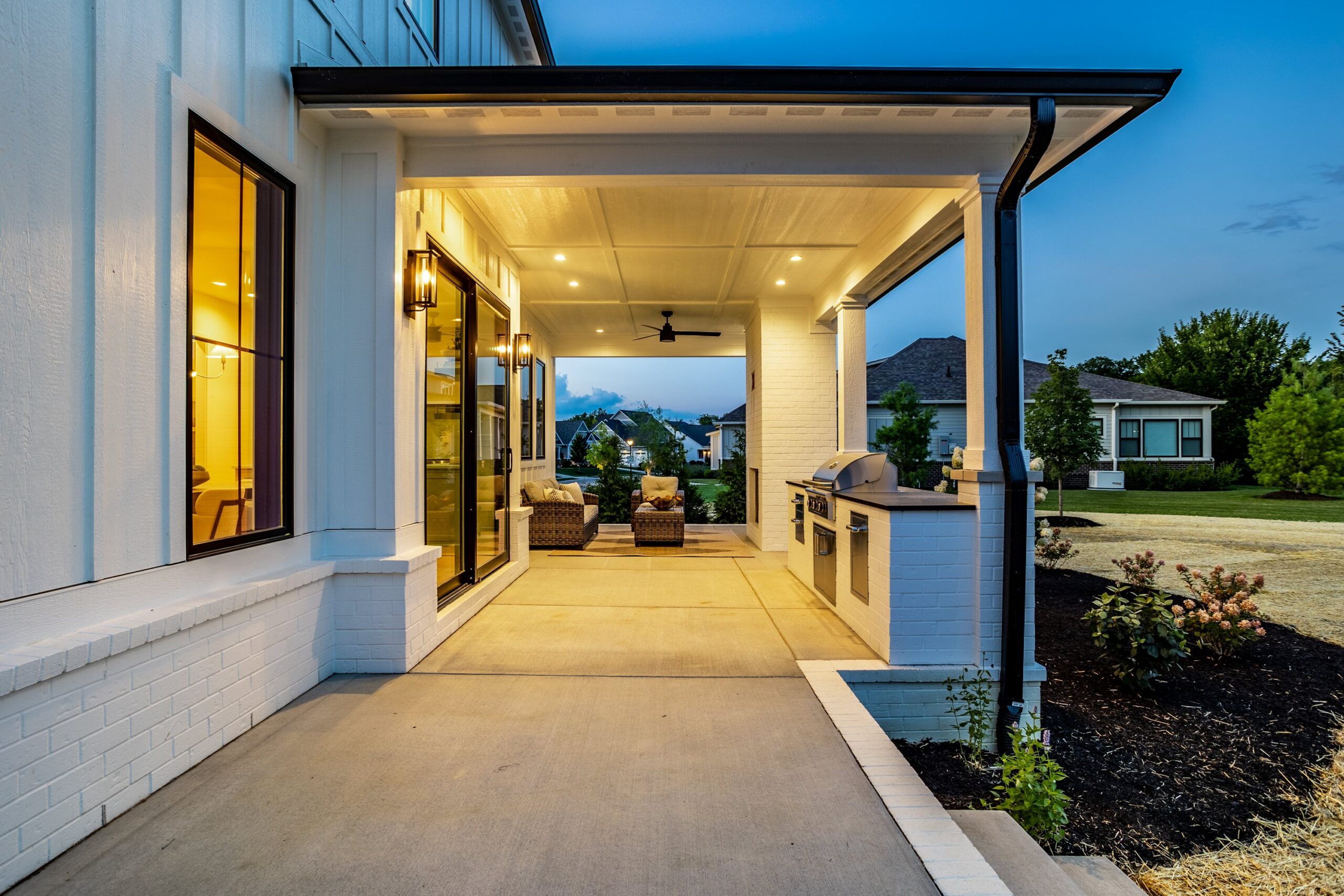 A modern covered patio at dusk, featuring outdoor seating and a built-in grill, with warm lighting and a view of a green lawn and neighboring houses.