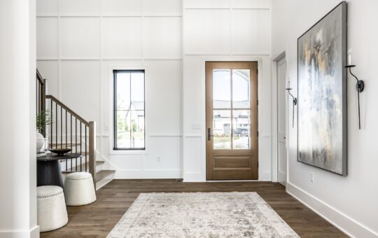 Modern entryway with light walls, wood flooring, a large area rug, a wooden front door with glass, a staircase with metal railing, a round table with stools, and abstract artwork on the wall. Natural light fills the space.