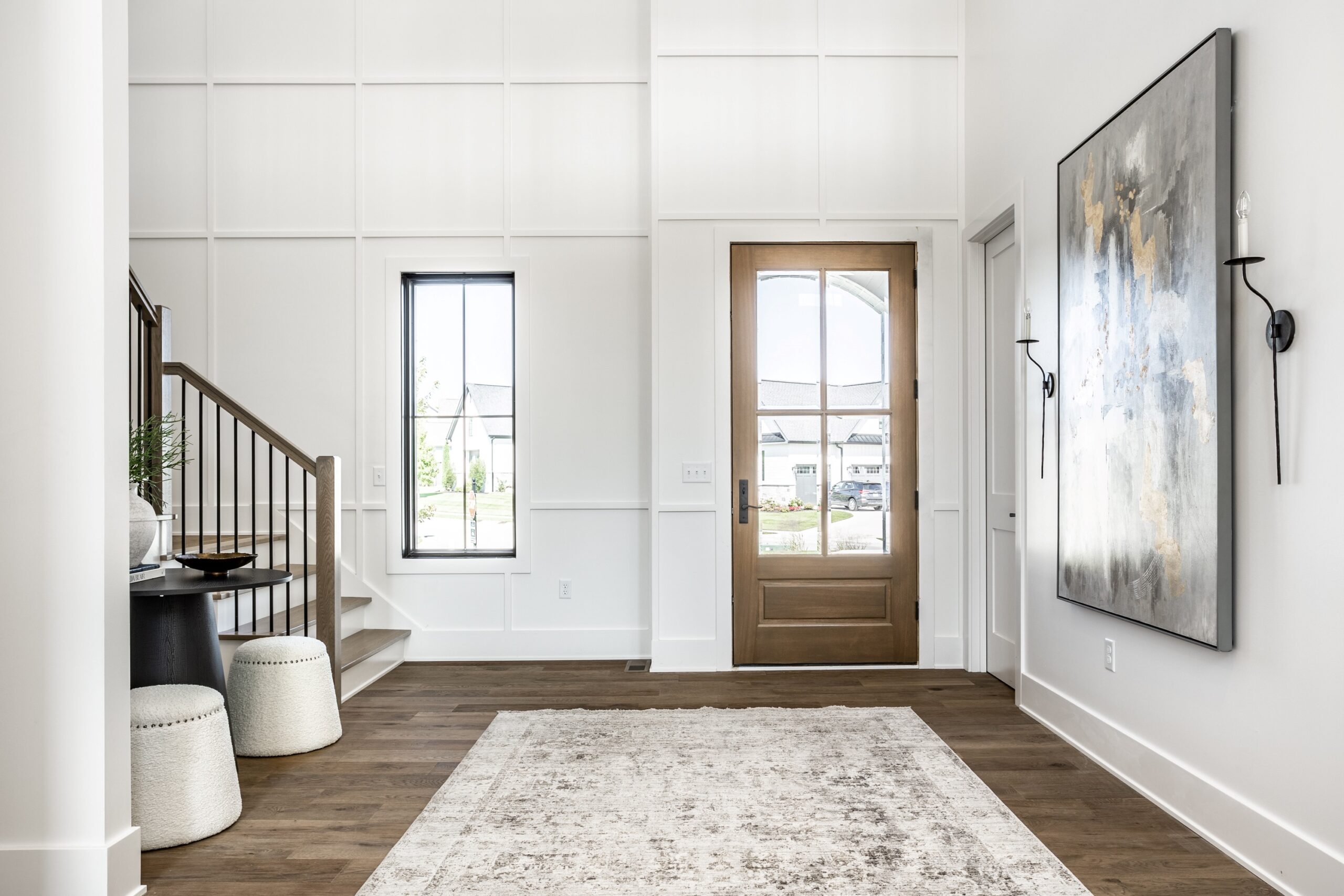 Modern entryway with light walls, wood flooring, a large area rug, a wooden front door with glass, a staircase with metal railing, a round table with stools, and abstract artwork on the wall. Natural light fills the space.