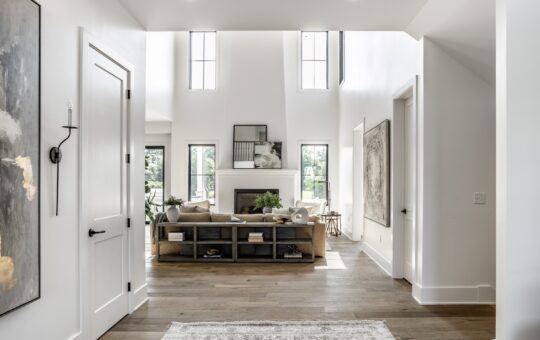 Bright, modern living room with tall windows, light wood flooring, neutral decor, and a beige sofa facing a fireplace. Large abstract art hangs on white walls; natural light fills the open space.