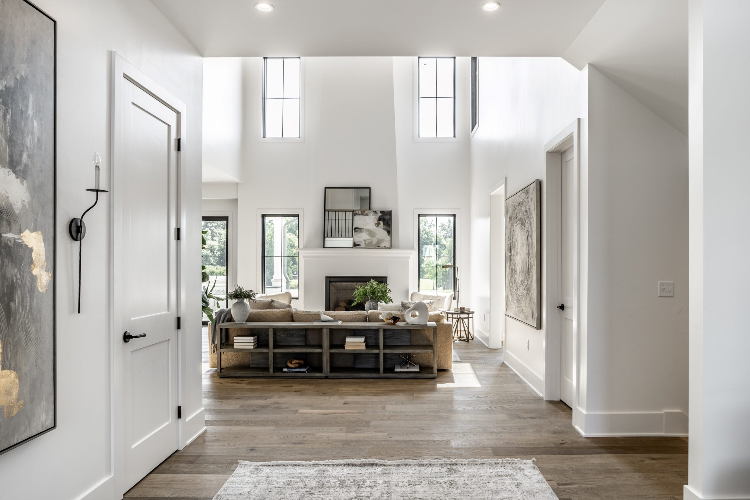 Bright, modern living room with tall windows, light wood flooring, neutral decor, and a beige sofa facing a fireplace. Large abstract art hangs on white walls; natural light fills the open space.