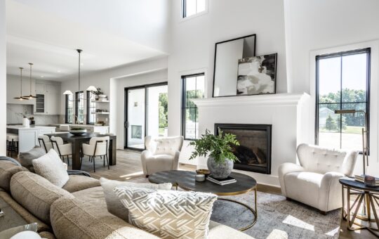 Bright, modern living room with large windows, neutral-toned furniture, a beige sofa, two armchairs, round coffee tables, a fireplace with abstract art, and an open view into a stylish kitchen.
