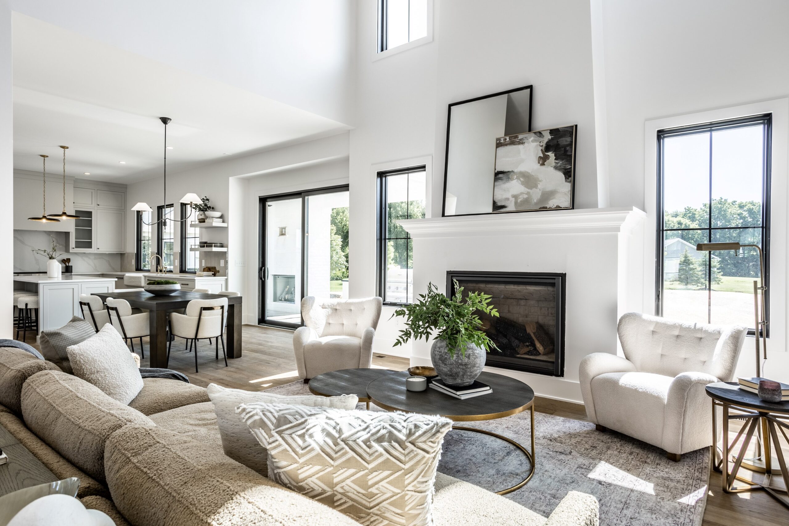Bright, modern living room with large windows, neutral-toned furniture, a beige sofa, two armchairs, round coffee tables, a fireplace with abstract art, and an open view into a stylish kitchen.