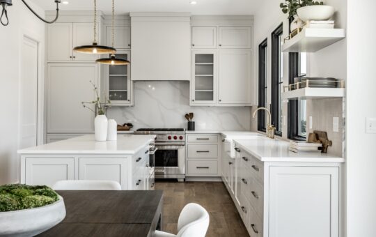 Modern kitchen with white cabinets, marble backsplash, gold fixtures, and open shelves with dishes and plants. A dark wood dining table with white chairs is in the foreground. Natural light streams through large windows.
