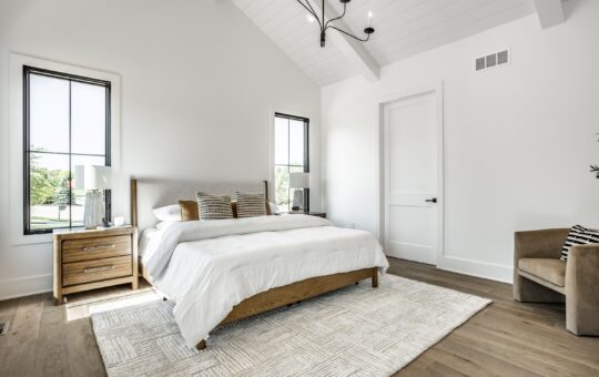 Bright, modern bedroom with white walls, vaulted ceiling, wooden floor, a large bed with white bedding, two wooden nightstands, a beige armchair, and windows letting in natural light.