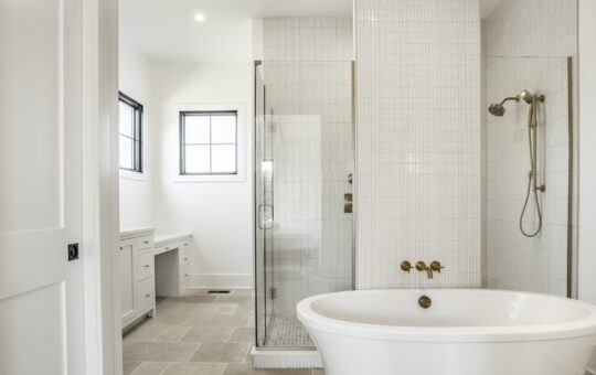 Modern bathroom with a freestanding white bathtub, glass-enclosed shower, gold fixtures, white tile walls, beige floor tiles, and a vanity with two windows providing natural light.