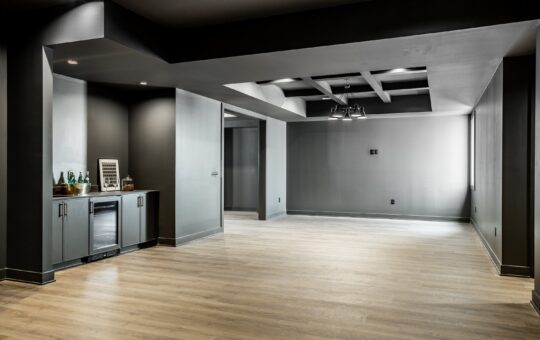 Modern, empty basement room with light wood flooring, dark gray walls and ceiling, built-in bar area with mini fridge and cabinets on the left, and ceiling lights throughout the space.