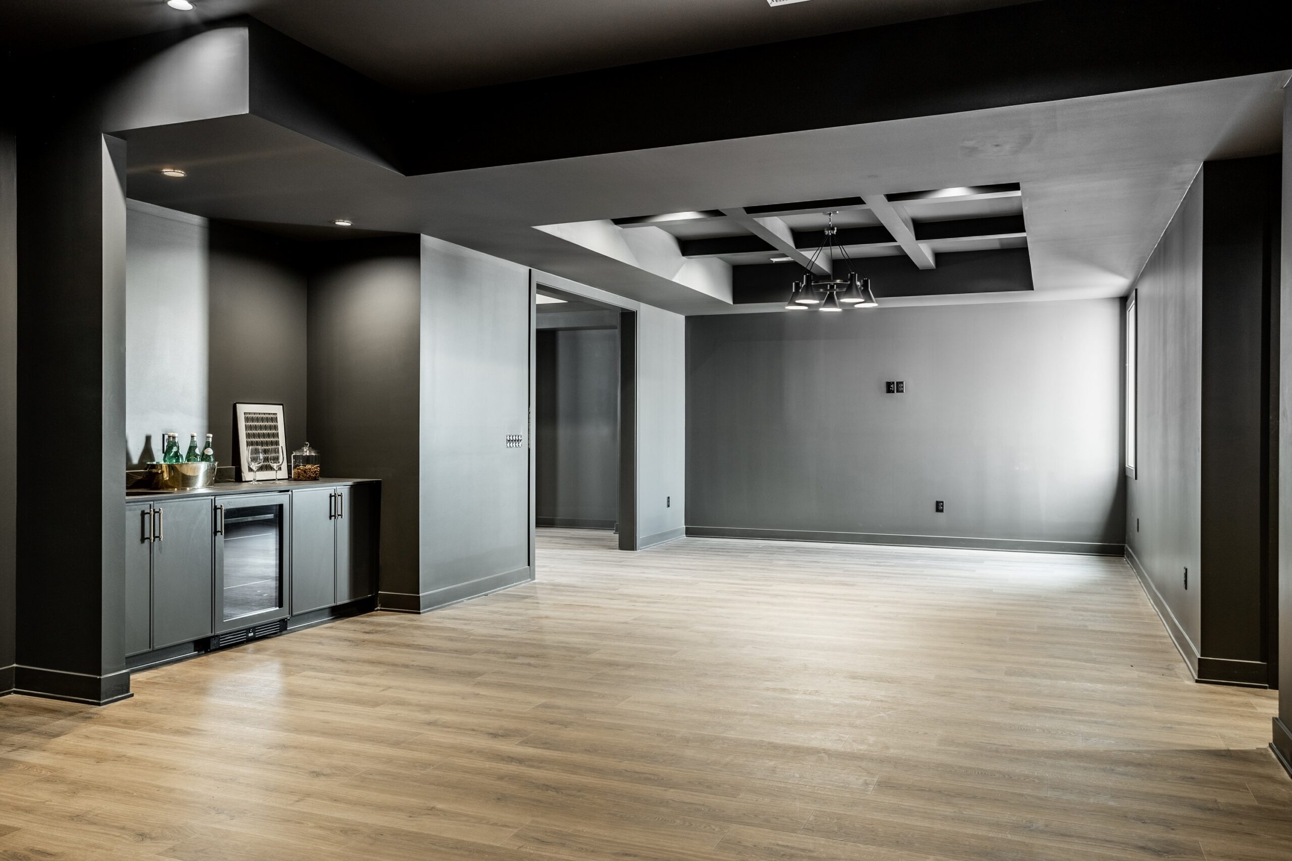 Modern, empty basement room with light wood flooring, dark gray walls and ceiling, built-in bar area with mini fridge and cabinets on the left, and ceiling lights throughout the space.