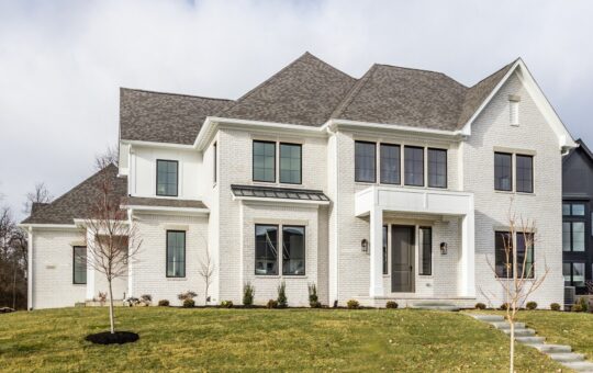 Two-story modern white brick house with large windows, gray shingle roof, covered front porch, and a landscaped front yard with young trees and shrubs on a cloudy day.