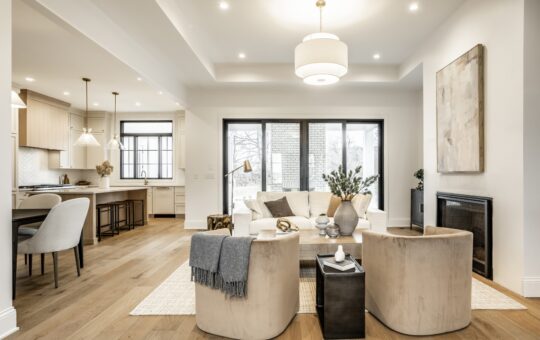 Modern open-concept living room and kitchen with light wood floors, neutral-toned furniture, a cozy seating area, large windows, pendant lighting, and contemporary decor.