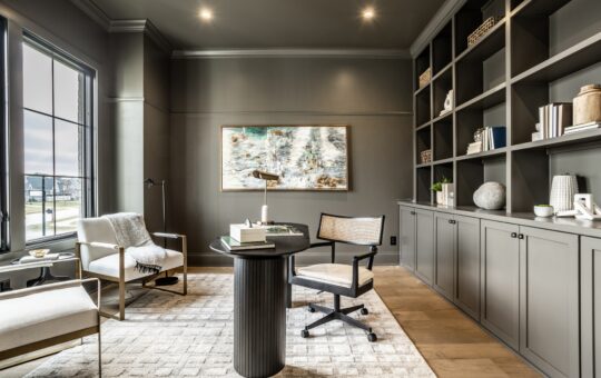 Modern home office with gray built-in shelves, desk and chair, two armchairs, a large window with outdoor view, abstract wall art, and decorative items neatly arranged on shelves. Neutral tones create a calm, elegant atmosphere.