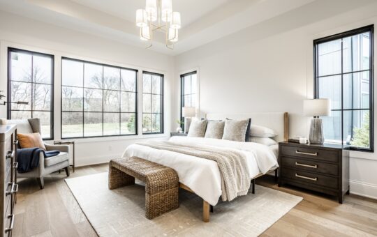 Bright modern bedroom with large windows, a bed with white bedding and neutral pillows, dark wood nightstands with lamps, a woven bench at the foot of the bed, and a cozy armchair by the window.
