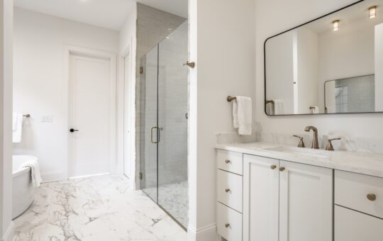 Modern bathroom with marble floor, glass shower, white cabinets, marble countertop, large mirror, and brushed metal fixtures. White walls and natural light create a clean, bright, and spacious atmosphere.