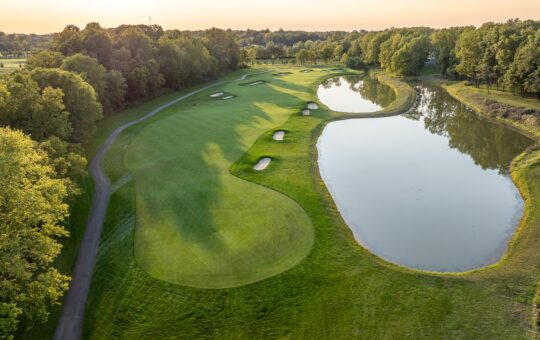 Aerial view of a golf course with a fairway, several sand bunkers, and a large pond on the right, surrounded by dense green trees under a clear sky at sunset.