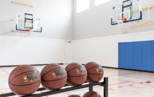 Four Spalding NBA basketballs rest on a rack in an indoor gym with two basketball hoops, blue wall padding, and large windows letting in natural light.