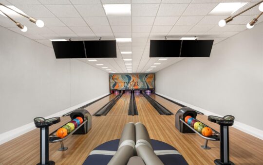 A modern bowling alley with four lanes, colorful bowling balls arranged on racks, overhead screens, and a mural at the end wall. The space is well-lit with white walls and a light wood floor.