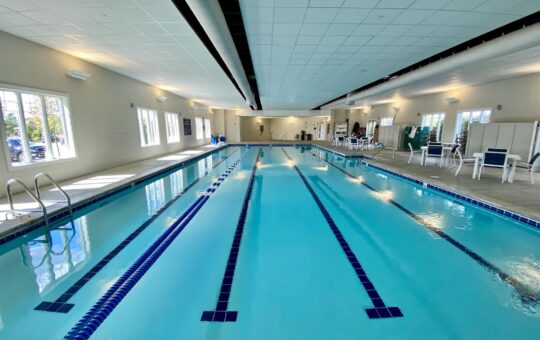 Indoor swimming pool with clear blue water and swim lanes; poolside chairs and tables are arranged along the walls under bright overhead lighting, with windows letting in natural light.