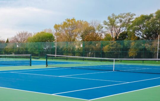 An empty outdoor tennis court with blue and green surfaces, surrounded by a wire fence. Trees with autumn foliage are visible in the background under a cloudy sky.