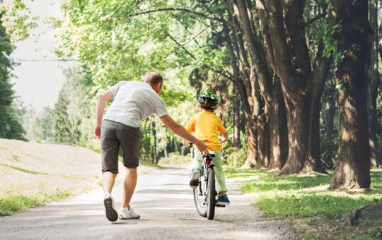 An adult helps a child ride a bicycle along a tree-lined path on a sunny day. The adult supports the child from behind, and both are surrounded by greenery and dappled sunlight.