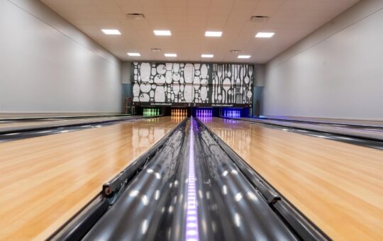 A close-up, low-angle view of multiple sleek, empty bowling lanes with polished wooden floors and colorful lights, leading toward a decorative black and white mural at the end of the alley.