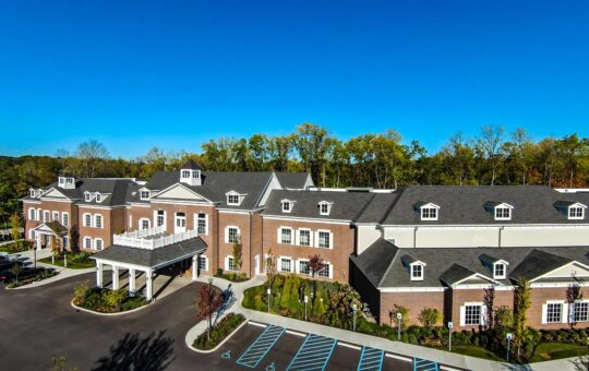 A large, modern brick building with white trim, surrounded by greenery and trees under a clear blue sky. There are parking spaces, including accessible spots, in front of the entrance with a covered driveway.