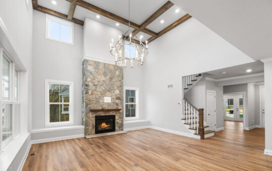 A spacious, modern living room with high ceilings, wood beam accents, a stone fireplace, large windows, wooden floors, and a grand chandelier. A staircase leads to the upper level. The room is bright and airy.