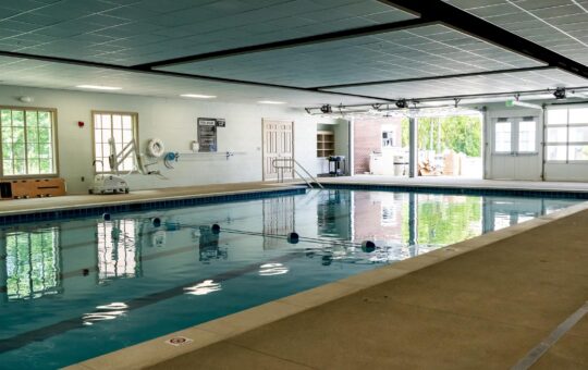 An indoor swimming pool with clear water, blue lane dividers, and a tiled deck. Large windows and glass doors let in natural light, and various pool equipment is visible along the white walls.