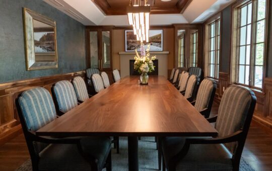 A long wooden conference table with twelve upholstered chairs in a formal meeting room, featuring large windows, framed artwork, wood paneling, and a vase of flowers centered on the table.