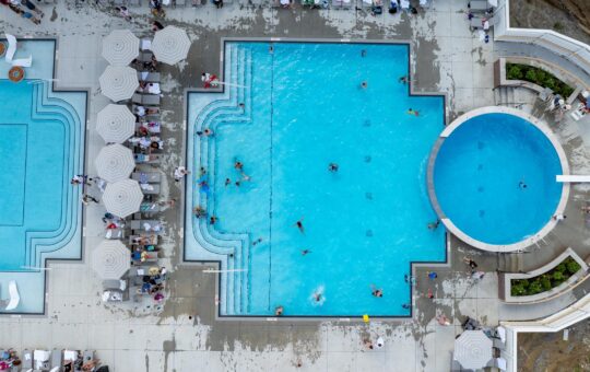Aerial view of a busy pool area with people swimming in a large rectangular pool, a smaller circular pool, and lounge chairs with umbrellas around the pools. Spectators are seated near the pools.