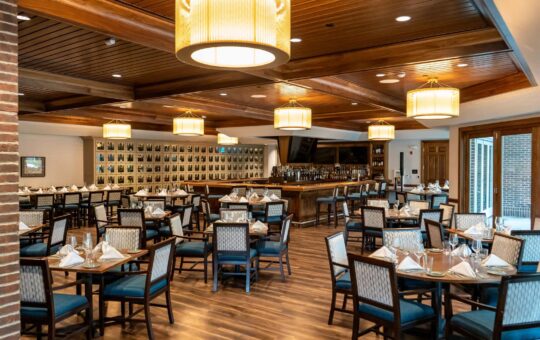 A spacious, modern restaurant dining room with wooden floors and ceiling, neatly arranged tables set with white napkins and glassware, blue cushioned chairs, large hanging pendant lights, and a bar in the background.