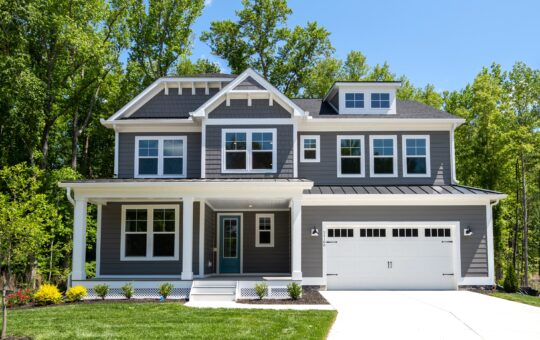 A modern two-story house with gray siding, white trim, a covered front porch, large windows, and a two-car garage, surrounded by a green lawn and trees in the background on a sunny day.