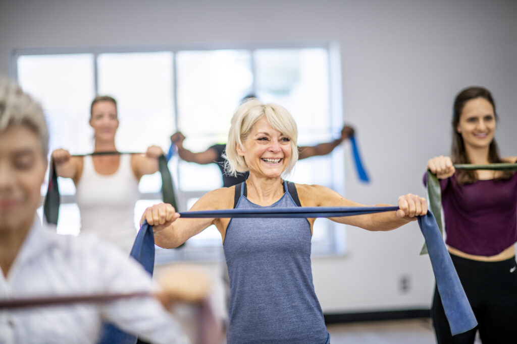 An elderly woman smiles as she works out during a fitness class. She is using an exercising band. The others around her are doing the same.