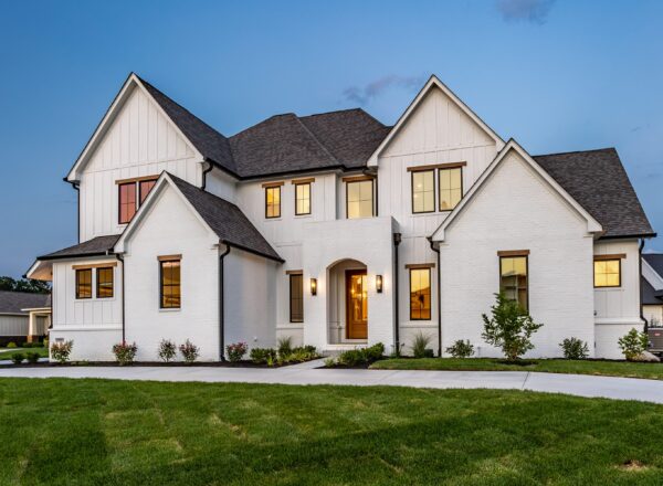 A modern two-story white brick house with large windows, a dark gray roof, and a well-kept lawn at dusk. Warm lights glow from inside, highlighting the entryway and front landscaping.